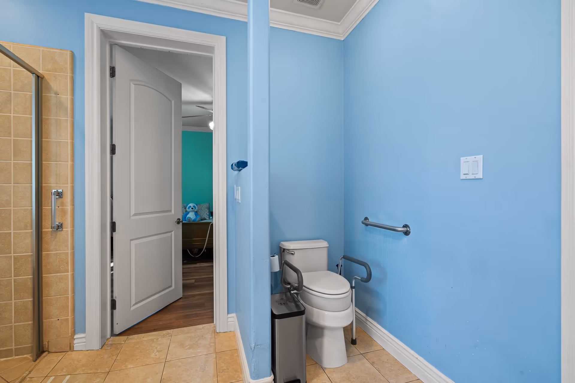 Bathroom with light blue walls, a white toilet equipped with safety rails, a trash can beside it, and a tiled shower area to the left. The bathroom door is open, revealing a room with teal walls, a ceiling fan, and a blue teddy bear on a piece of furniture.