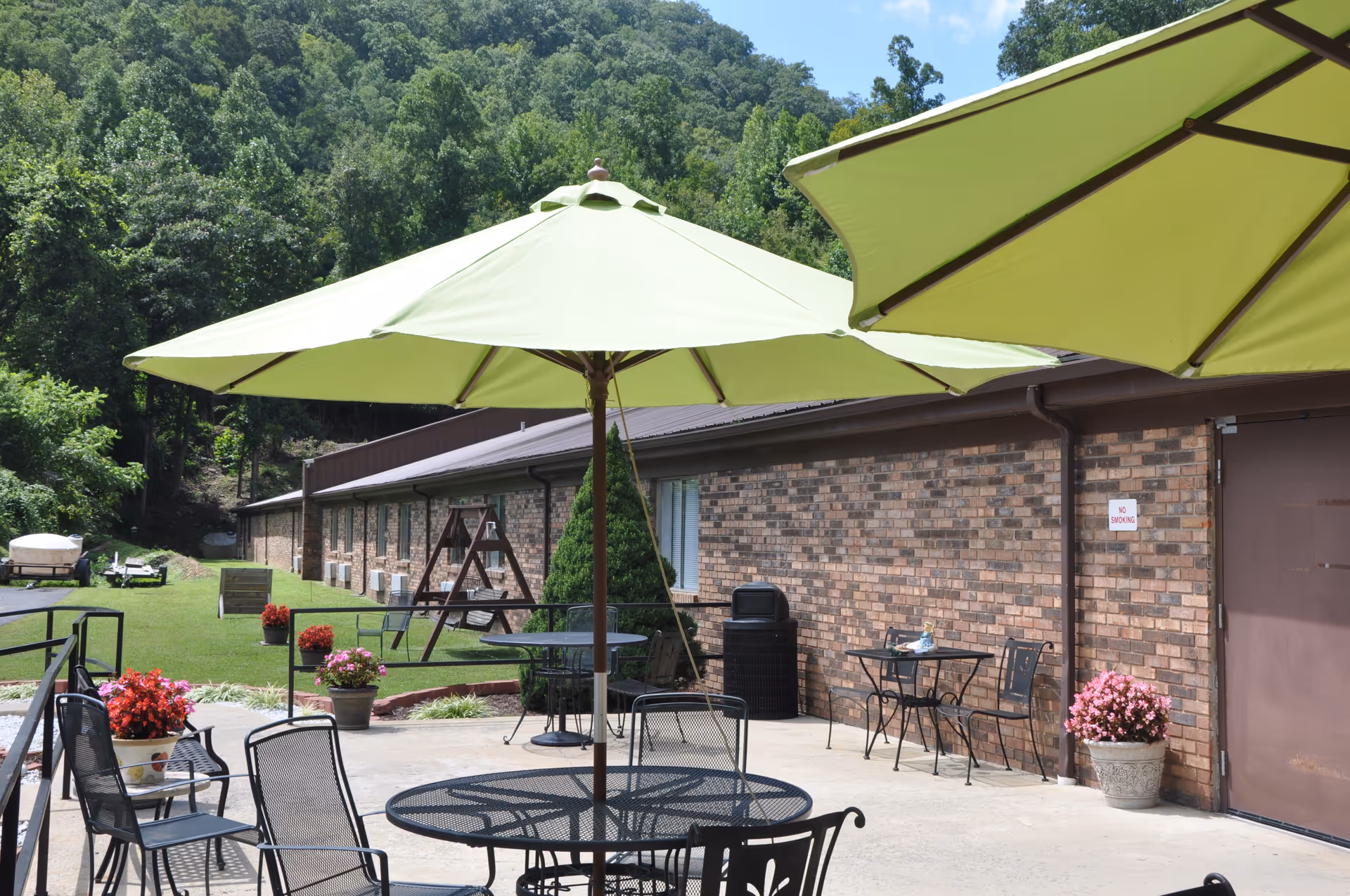 Outdoor patio area with black metal tables and chairs under large green umbrellas. There are potted flowers and a brick building wall in the background, with a grassy area and trees behind it.