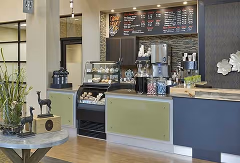 Interior view of a coffee shop counter area with a menu board displaying various drink options. The counter has a display case with pastries, coffee machines, and jars of candy. Decorative items including small animal figurines and plants are placed on a nearby round table.