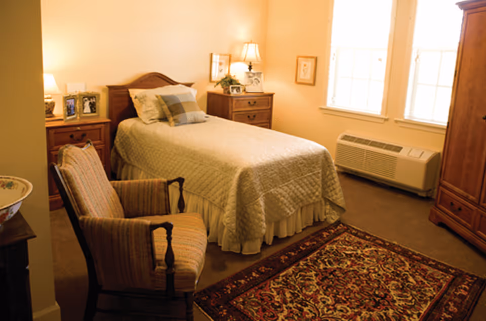 Cozy single bedroom with a made bed, upholstered armchair, wooden nightstands, dresser and a patterned area rug.