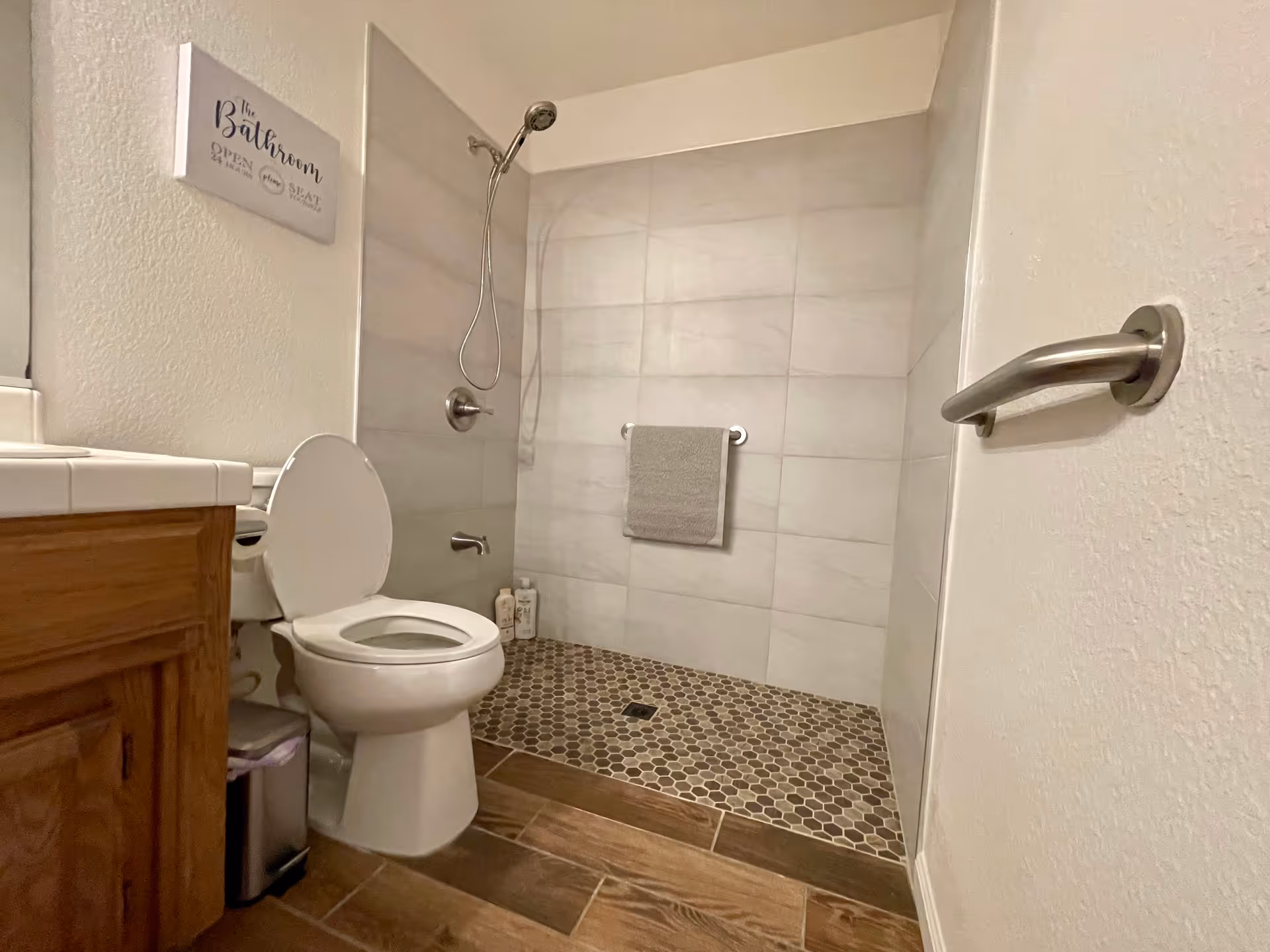 A bathroom with a toilet, a wooden vanity with a white countertop, and a walk-in shower with a handheld showerhead and a towel hanging on a bar. The shower floor has small hexagonal tiles, and the bathroom floor has wood-like tiles. A wall sign above the toilet reads 'The Bathroom Open When Seat Down'.