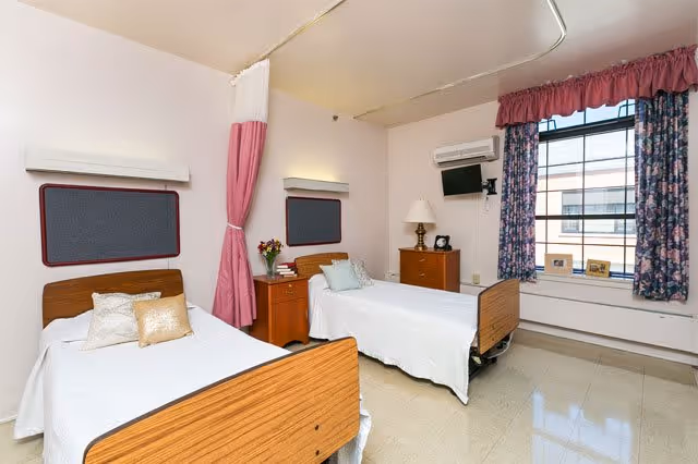 A bright and clean room with two single beds, each with white bedding and pillows. Between the beds is a small wooden nightstand with a vase of flowers and some books. The room has a large window with floral curtains, a wall-mounted TV, a lamp on a wooden dresser, and a pink privacy curtain hanging from the ceiling.