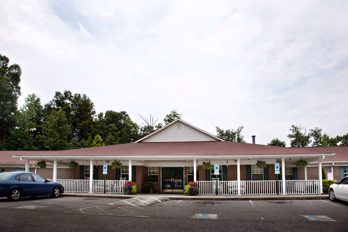 Front exterior view of a single-story brick building with a red roof and white railing porch. There are hanging plants and flower pots near the entrance. Several handicap parking spaces are visible in front of the building, along with two parked cars on either side.