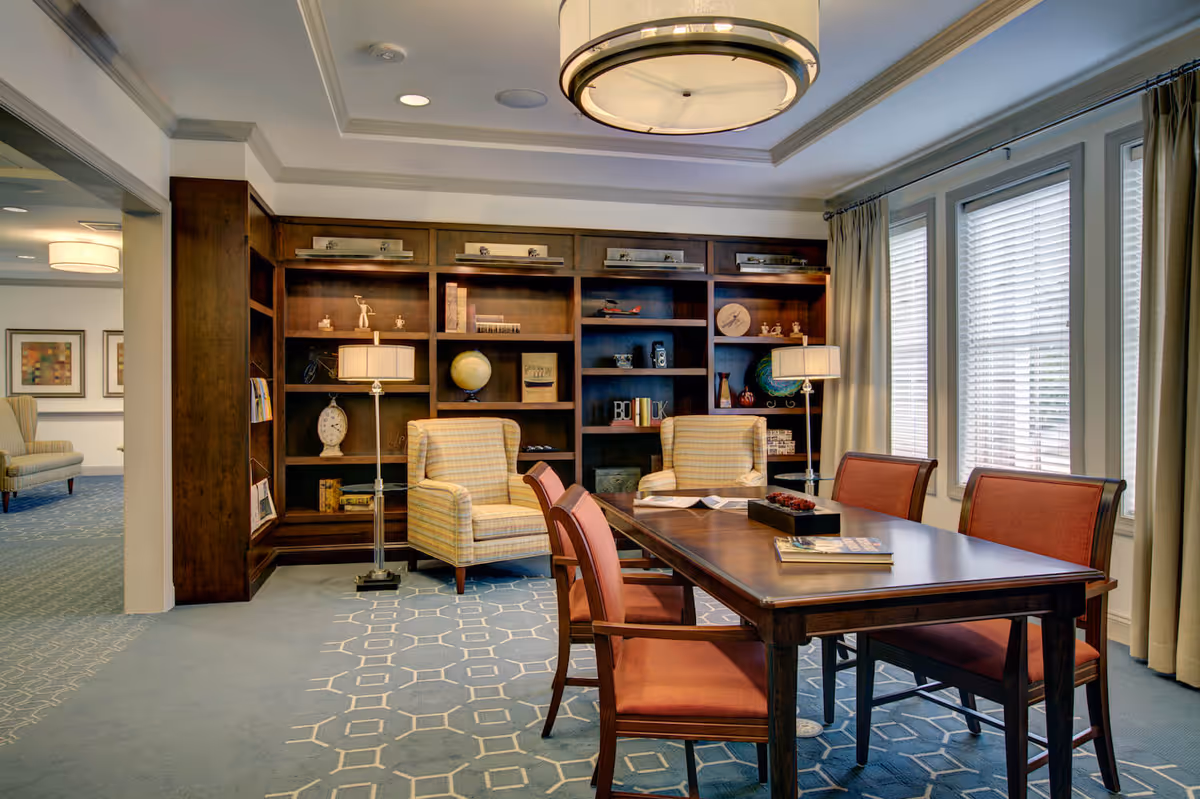 A bright senior living common room with a wooden table and chairs, armchairs, built-in bookshelves, and large windows with blinds.