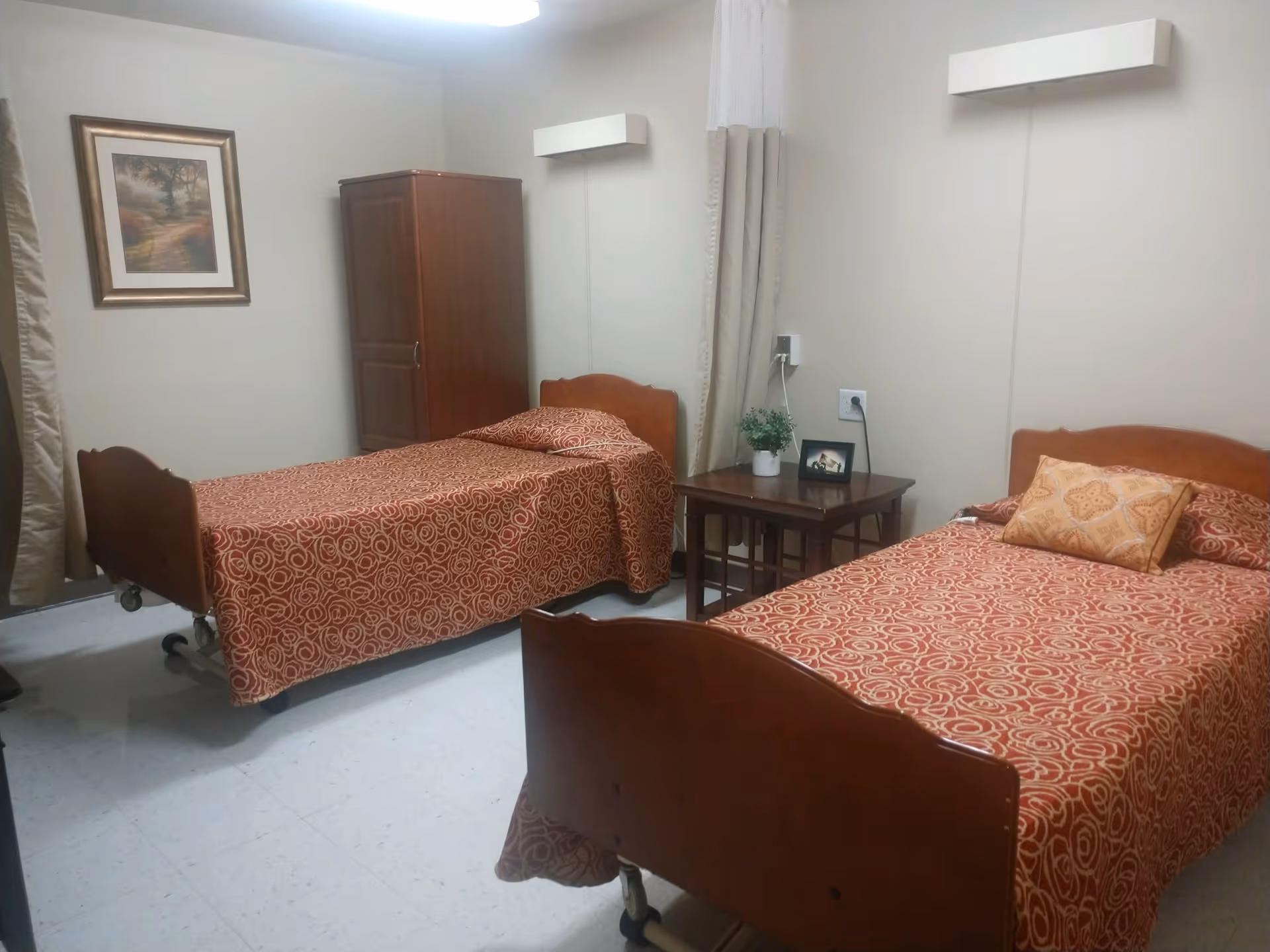 A nursing facility bedroom with two twin hospital-style beds with patterned bedspreads, a nightstand, wardrobe, and wall art.