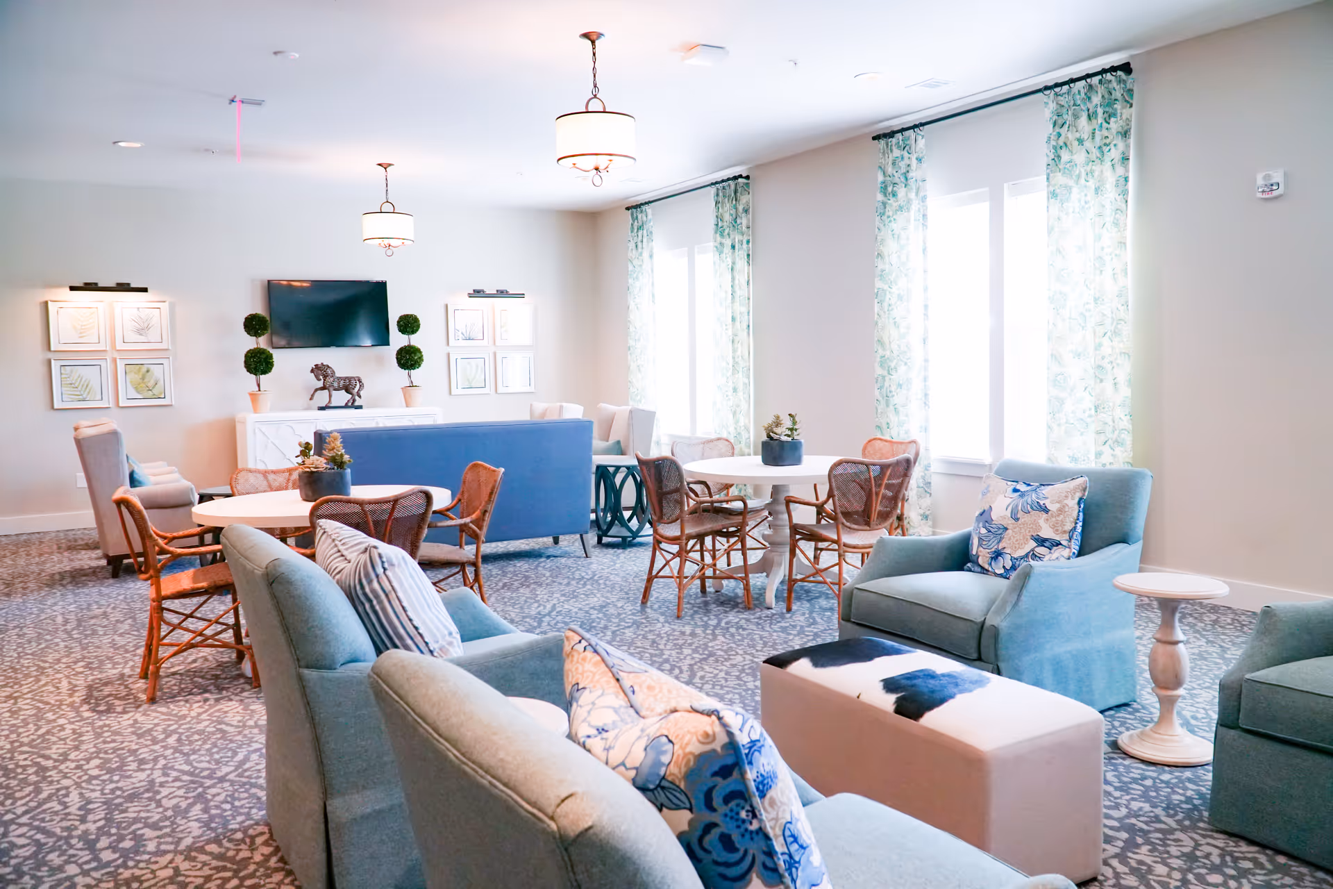 A bright and spacious living room area with multiple seating options including blue upholstered armchairs, a blue sofa, and wooden chairs around round tables. The room features patterned carpet, floral curtains on large windows, framed botanical artwork on the walls, and a flat-screen TV mounted above a white console table with decorative plants and a horse statue.