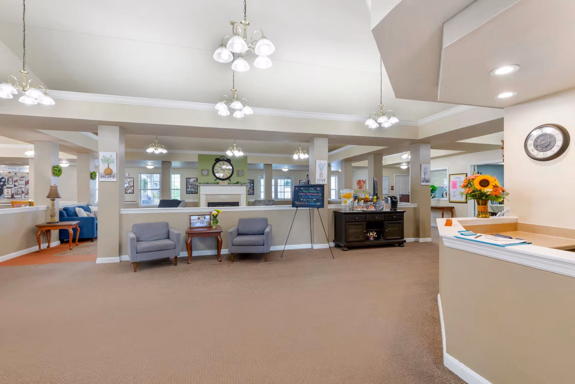 Open senior-living common area with lounge chairs, a reception desk to the right, and a fireplace with seating in the background.