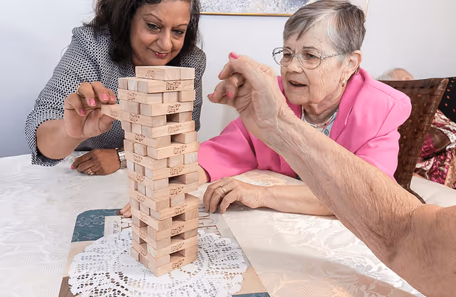 Two women, one elderly and one younger, are playing a game of Jenga at a table covered with a white lace tablecloth. The elderly woman is wearing glasses and a pink jacket, while the younger woman is smiling and carefully removing a Jenga block. A third person's hand is also visible reaching toward the tower.