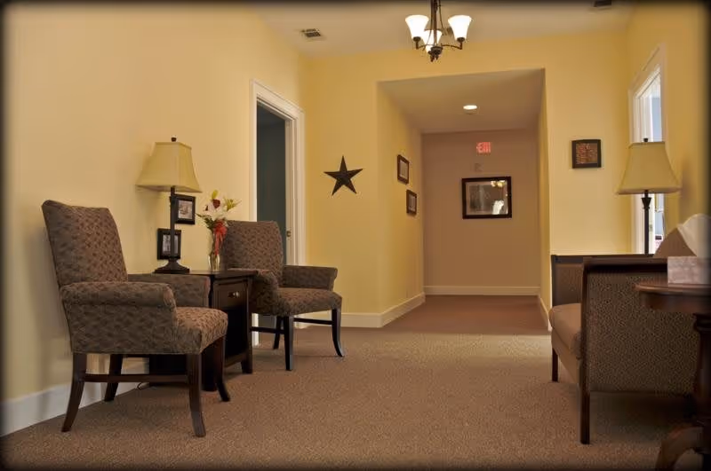 Carpeted hallway seating area with upholstered chairs, side tables, lamps, and framed wall decor under warm yellow walls.