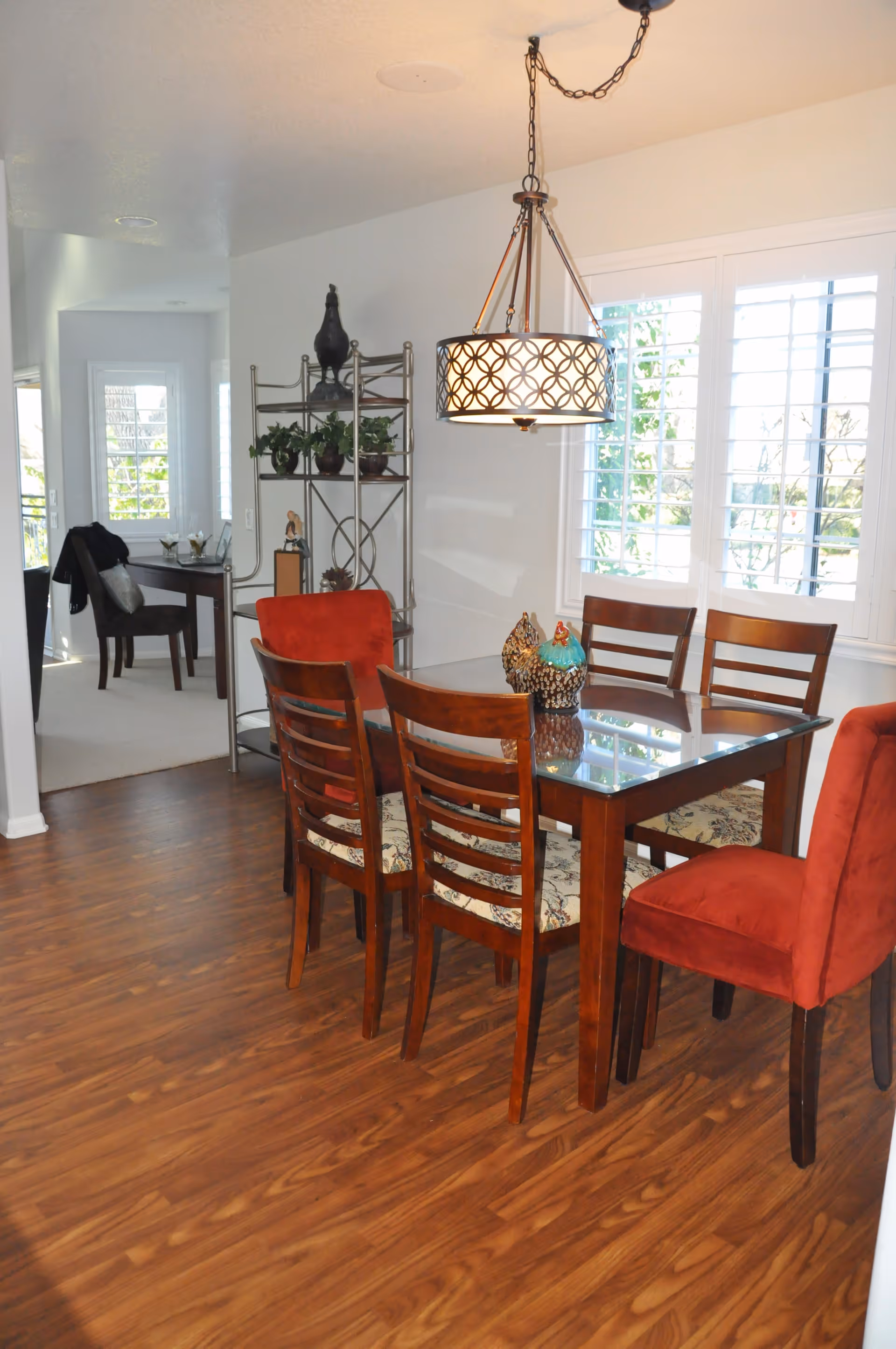 A dining room with a glass-top wooden table surrounded by six chairs, two of which have red upholstery. A decorative light fixture hangs above the table. There is a metal shelving unit with plants and decorative items against the wall, and a window with white shutters letting in natural light. In the background, there is a small desk with a chair and a jacket draped over it.