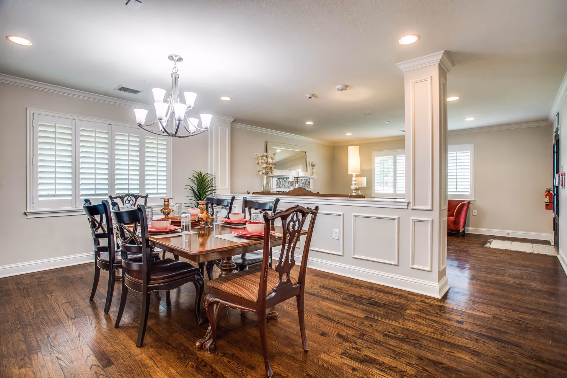 A well-lit dining room with a wooden dining table set for six people. The table has red plates, bowls, and glasses arranged on beige placemats. There are six dark wooden chairs around the table. The room features hardwood flooring, white walls with decorative paneling, a chandelier above the table, and windows with white plantation shutters. In the background, there is a partial view of a living area with a lamp and a red armchair.
