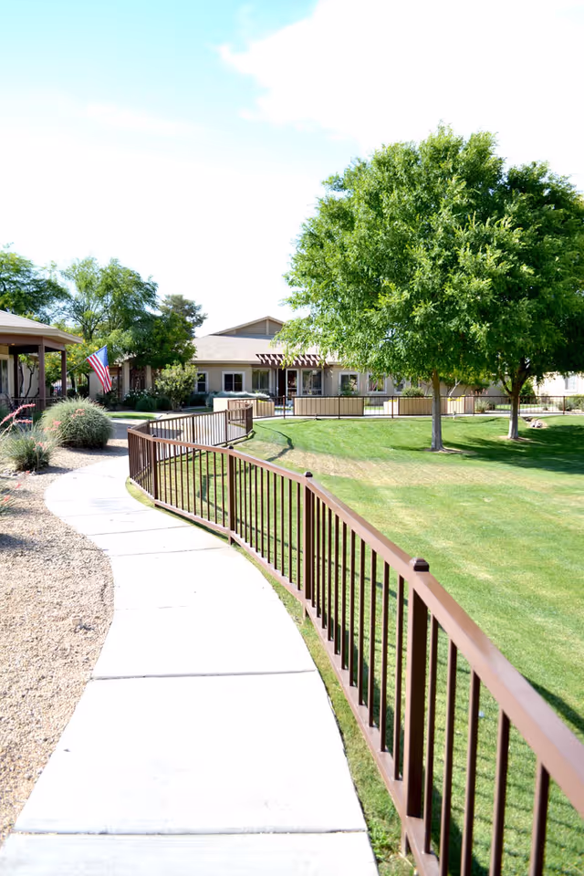 Curved concrete sidewalk bordered by a brown metal railing leading through a landscaped outdoor area with green grass, trees, shrubs, and a building in the background under a partly cloudy sky.