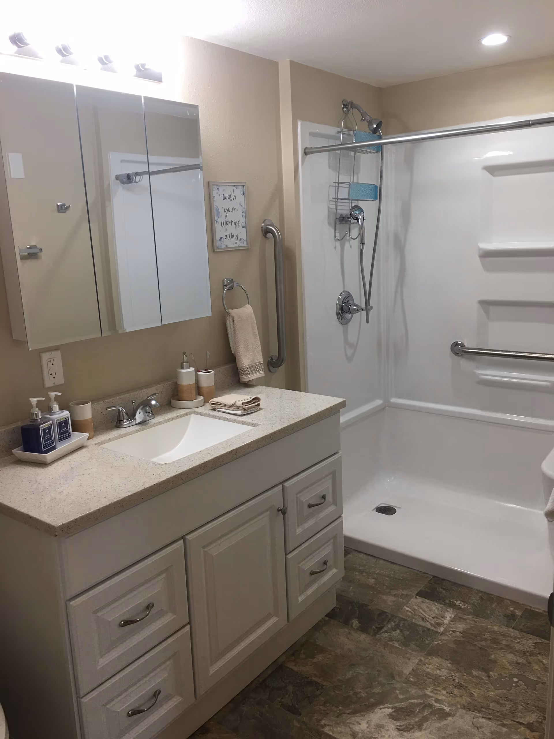 A clean and modern bathroom featuring a white vanity with a sink, a large mirror with three panels above it, and a walk-in shower with a sliding glass door and grab bars. The countertop has soap dispensers, a toothbrush holder, and neatly folded towels. The floor has a stone tile pattern, and the walls are painted beige.