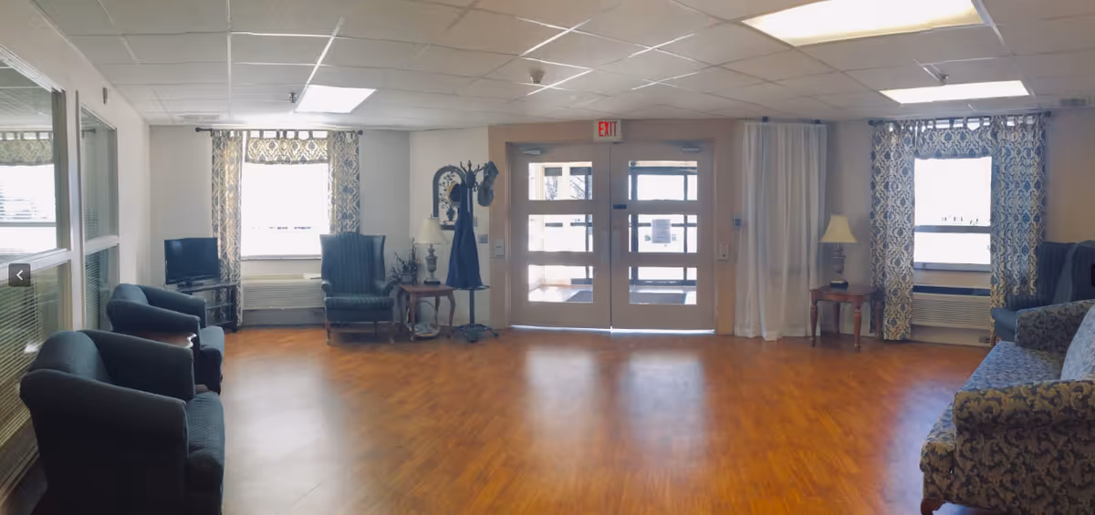 A spacious common area with wooden flooring, multiple upholstered chairs and sofas arranged around the room, two windows with patterned curtains, a small TV on a stand, a coat rack, two side tables with lamps, and double glass doors leading outside.