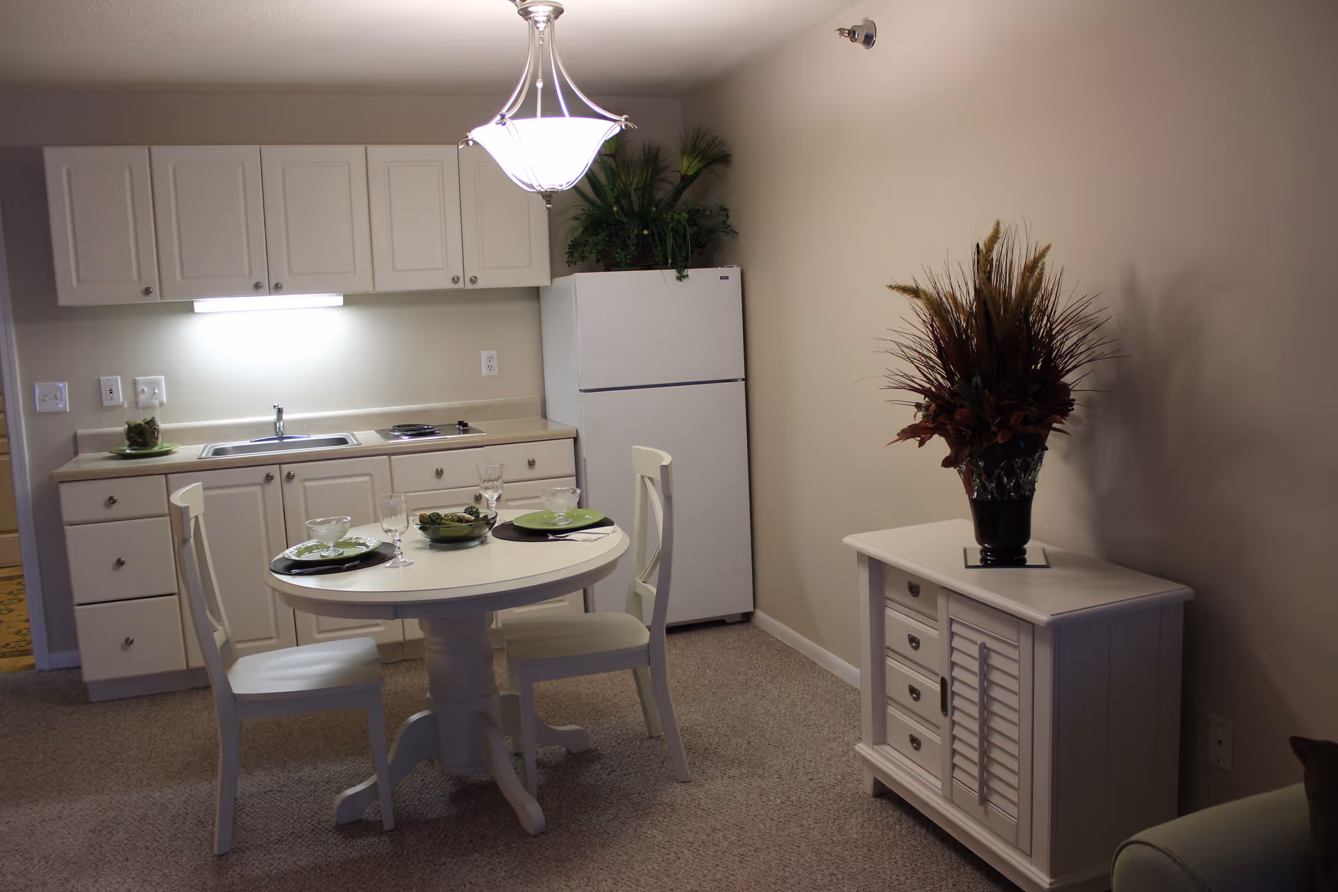 A small kitchen and dining area in a senior living facility. The kitchen has white cabinets, a countertop with a sink, a stovetop, and a white refrigerator. In front of the kitchen is a round white dining table set for two with plates, bowls, and glasses. To the right, there is a white cabinet with a decorative vase containing dried plants. The room is softly lit by a ceiling light fixture.
