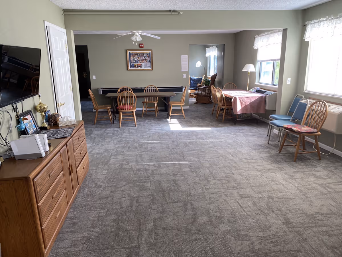 A spacious common area with gray carpet and light green walls. There are several wooden chairs and tables, including a table with a pink tablecloth near two windows with white curtains. A wooden dresser with a TV mounted above it is on the left side, along with some framed photos and decorative items. A ceiling fan with lights is mounted on the ceiling, and a cozy seating area with a rocking chair and cushions is visible through an open doorway.