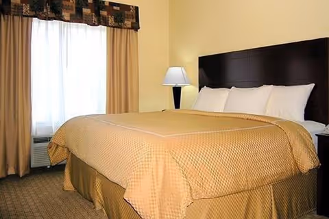 A bedroom with a large bed covered in a beige quilt and matching bed skirt. The bed has four white pillows and a dark wooden headboard. Next to the bed is a nightstand with a lamp. A window with beige curtains and a patterned valance is visible on the left side of the room.