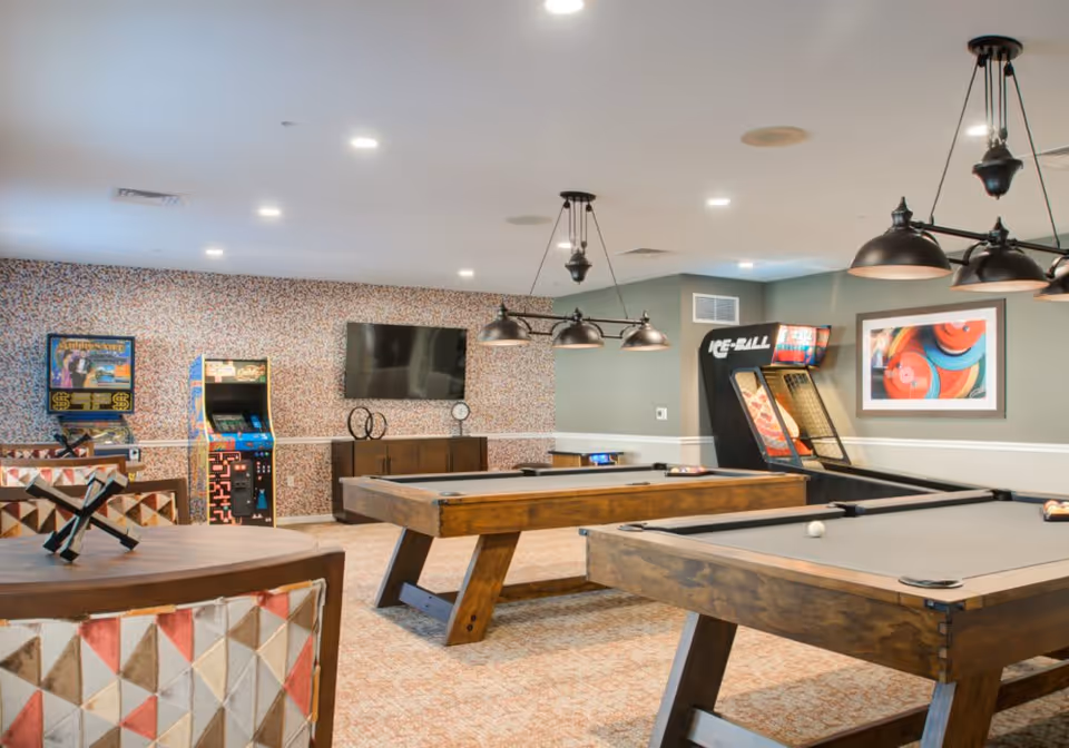 A recreational room featuring two wooden pool tables with overhead lighting, arcade game machines against the wall, a flat-screen TV mounted on a patterned wallpapered wall, and a framed colorful abstract artwork on a gray wall.