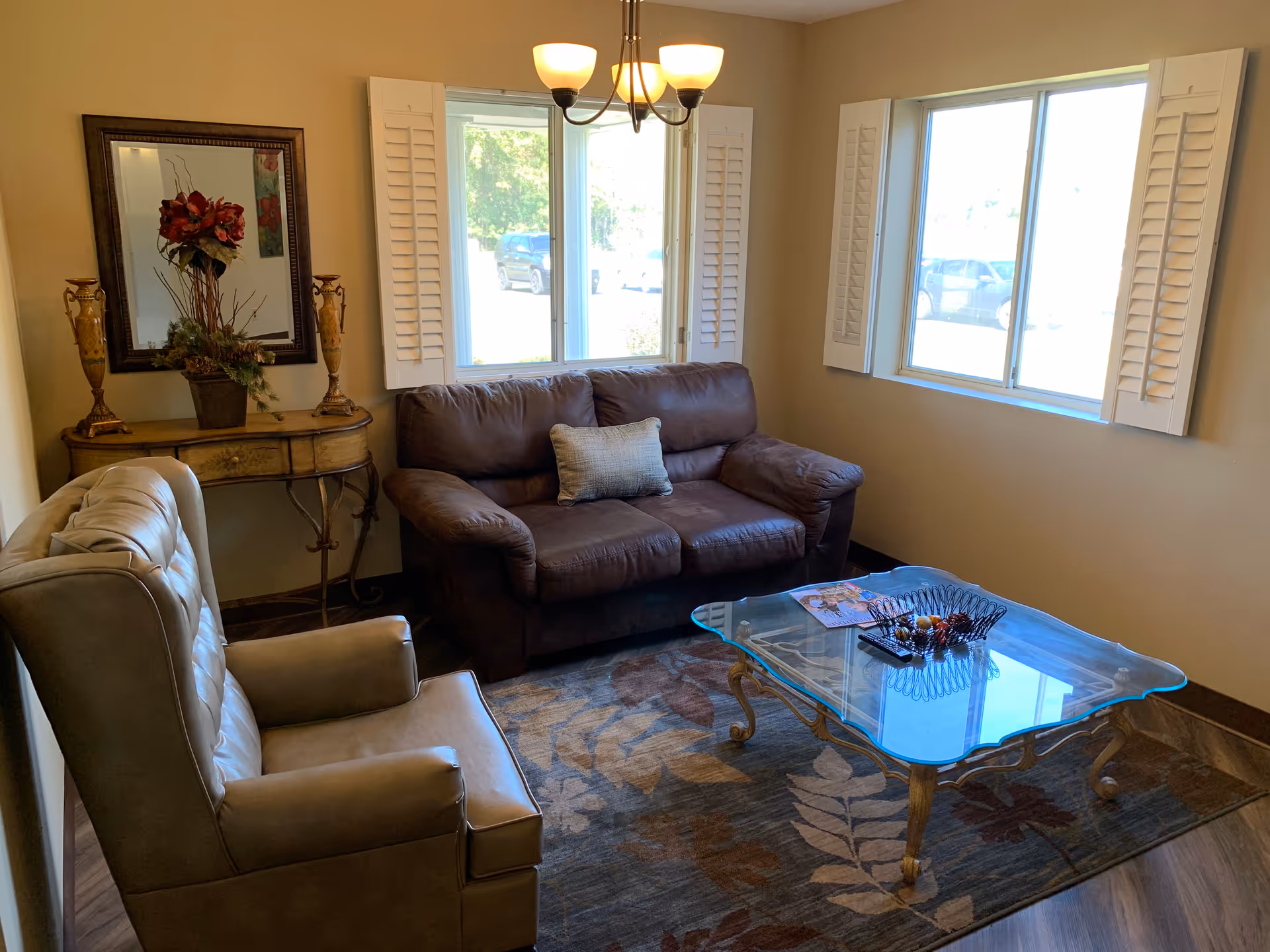 A cozy living room with a brown leather loveseat, a beige armchair, and a glass coffee table on a patterned rug. There is a wooden console table with decorative vases and a floral arrangement beneath a framed mirror. Two windows with white shutters let in natural light.
