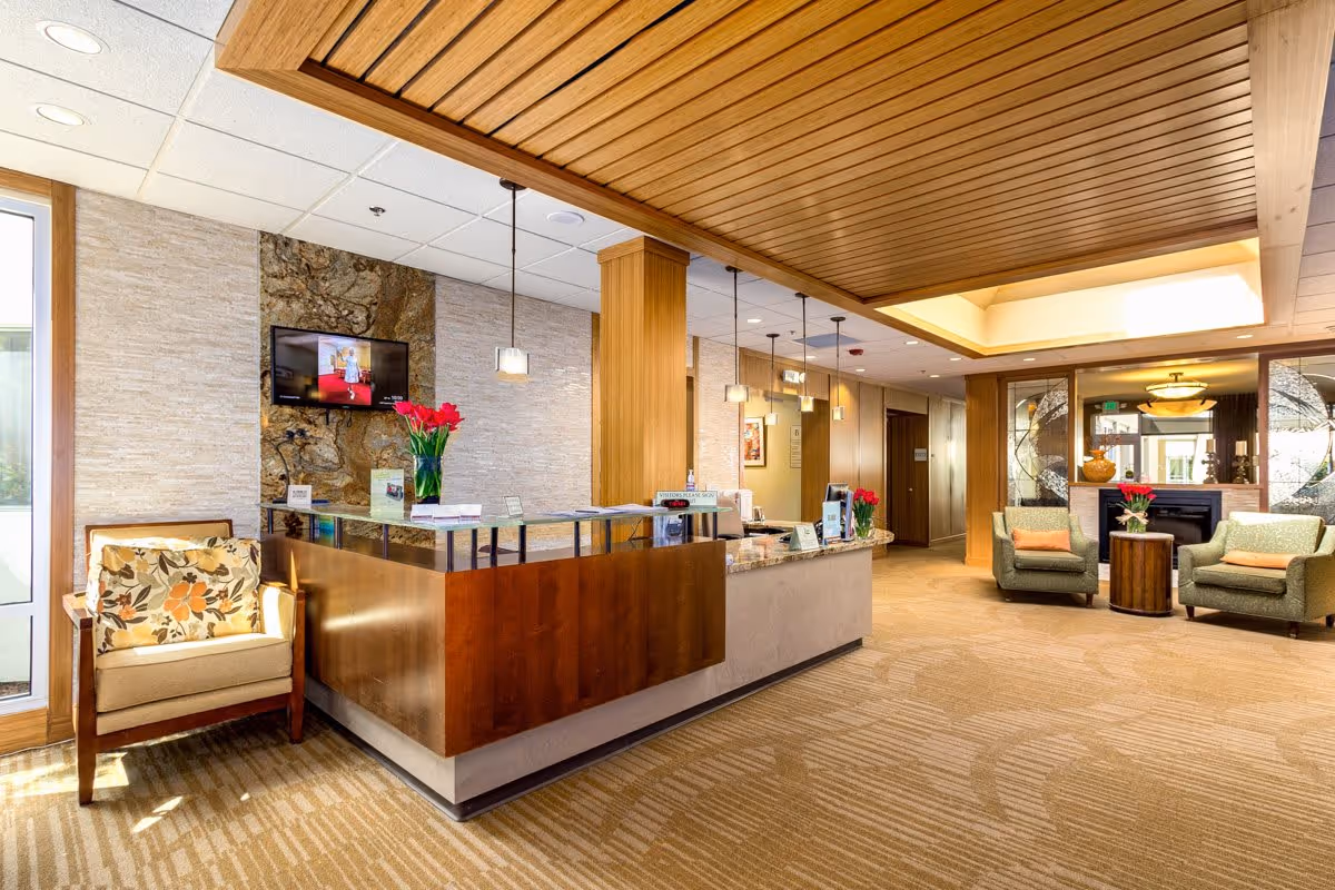 A bright and welcoming reception area in a senior living facility named CRISTA, featuring a wooden reception desk with a granite countertop, a vase with red flowers, a TV mounted on a stone accent wall, comfortable armchairs with cushions, pendant lighting, and a carpeted floor with a patterned design.