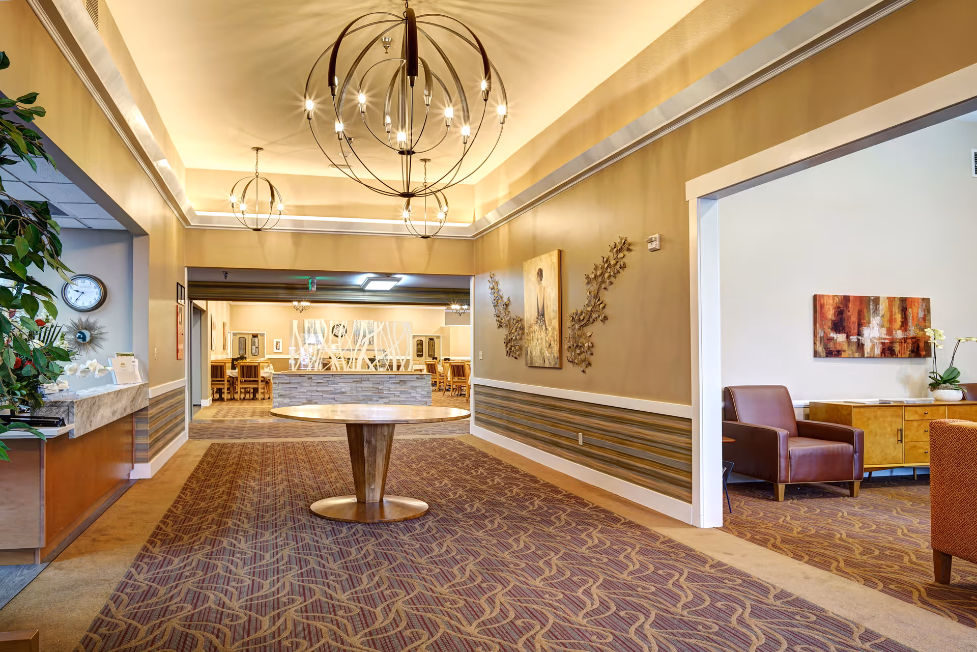 Interior view of a senior living facility hallway with a round wooden table in the center, modern spherical chandeliers hanging from the ceiling, and a seating area with chairs and artwork on the walls to the right. The hallway leads to a dining area with tables and chairs visible in the background.