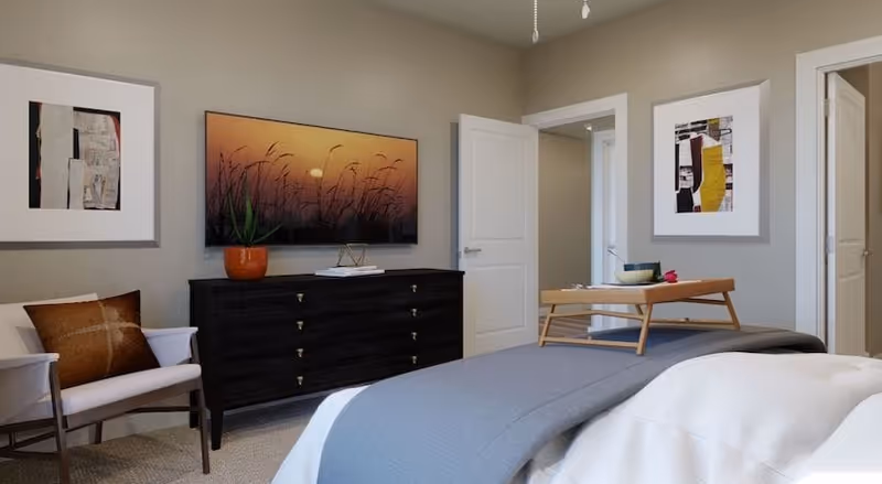 A bedroom with a bed covered in white and gray bedding, a wooden tray with a bowl and a rose on the bed, a dark wooden dresser with a potted plant and a framed picture of a sunset above it, a white armchair with a brown pillow, and two open doors leading to other rooms. The walls are light gray and decorated with framed abstract artwork.