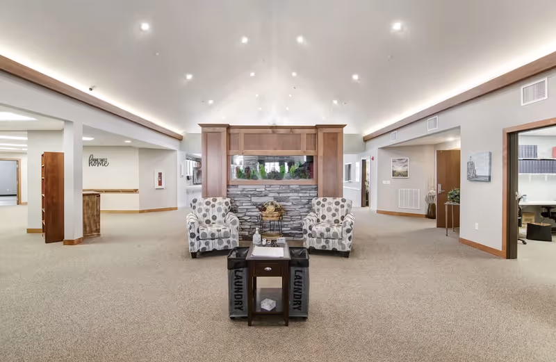 A spacious senior living facility common area with beige carpeting and a high ceiling with recessed lighting. In the center, there are two patterned armchairs facing a small wooden table with two laundry baskets on either side. Behind the chairs is a stone fireplace with a wooden mantel and a built-in aquarium. The walls are light-colored with wooden trim, and there are open doorways leading to other rooms. A wall decoration reads 'Home Sweet Home'.