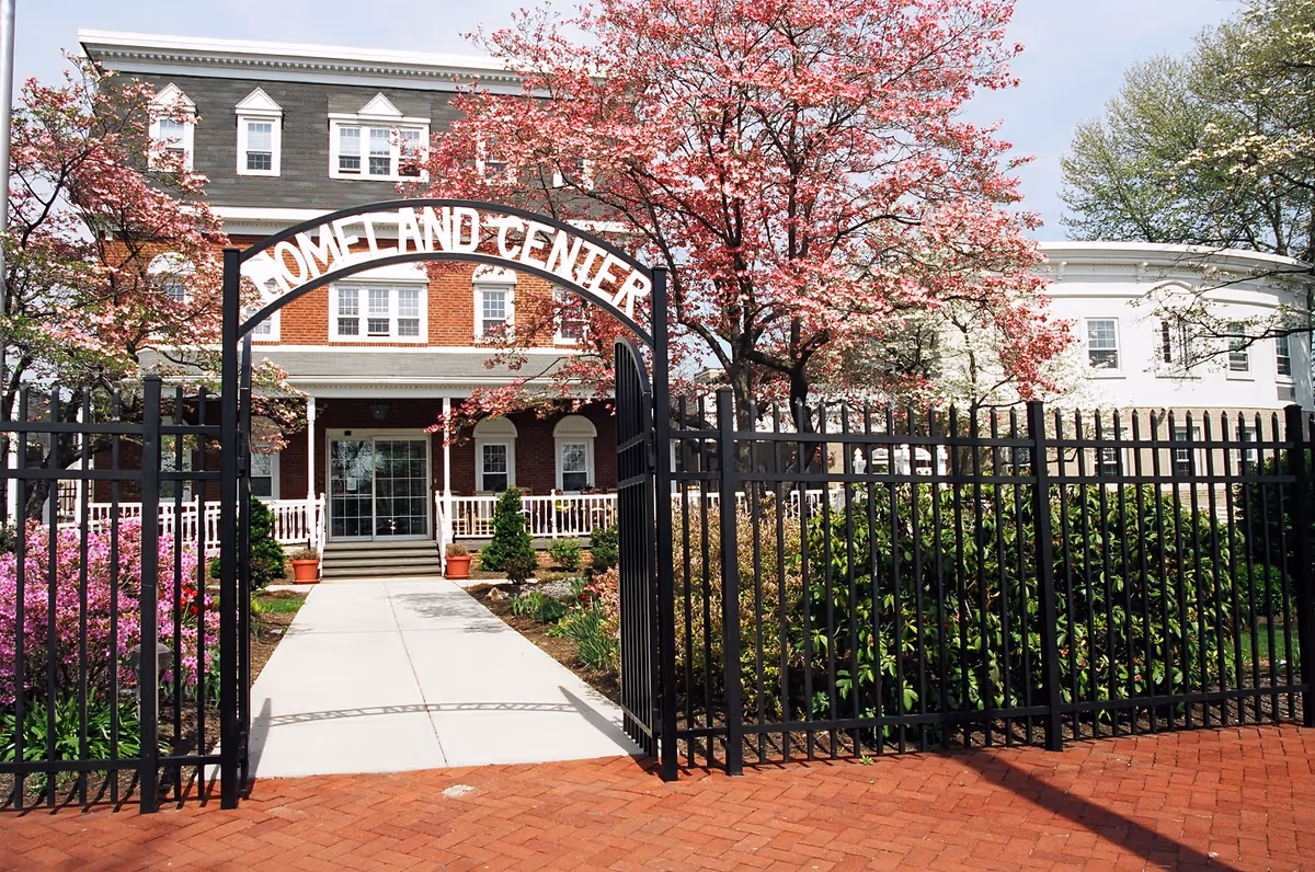 Entrance gate with an arch reading 'Homeland Center' leading to a brick building with white trim. The pathway is flanked by blooming pink and white flowering trees and green shrubs, with a black metal fence surrounding the garden area.
