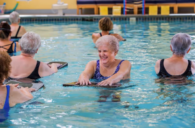 A group of elderly women participating in a water exercise class in an indoor swimming pool, holding kickboards and smiling.