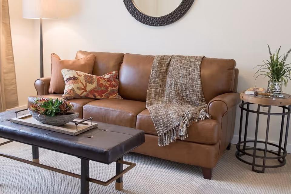 A cozy living room area featuring a brown leather sofa adorned with a patterned throw pillow and a textured blanket. In front of the sofa is a black leather ottoman-style coffee table with a decorative tray holding a bowl of succulents. To the right of the sofa is a round wooden side table with a small plant and some books. A floor lamp with a white shade stands to the left, and a round decorative mirror hangs on the wall above the sofa.