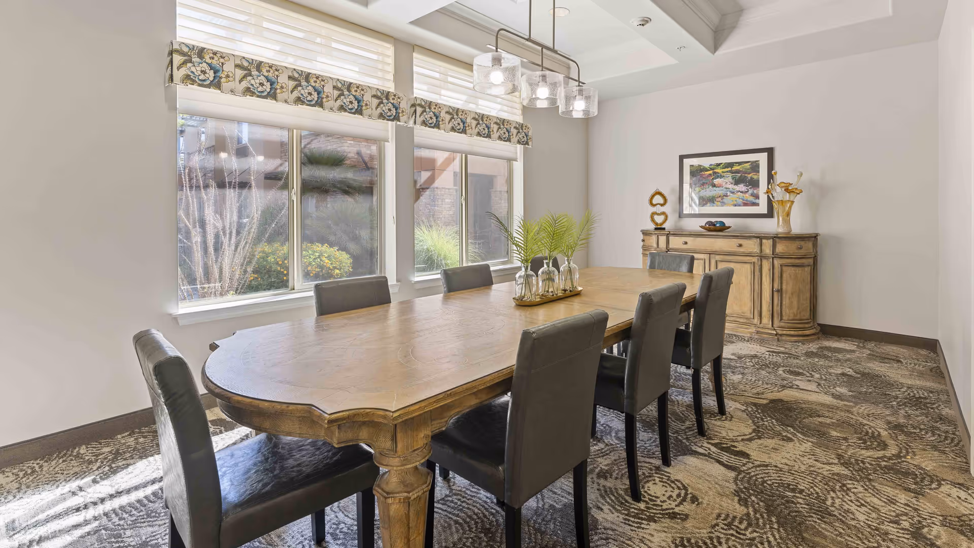 A bright dining room with a large wooden table surrounded by six black leather chairs. The room features three large windows with floral valances, a patterned carpet, a wooden sideboard with decorative items, and a framed painting on the wall. A modern light fixture with three glass shades hangs above the table.
