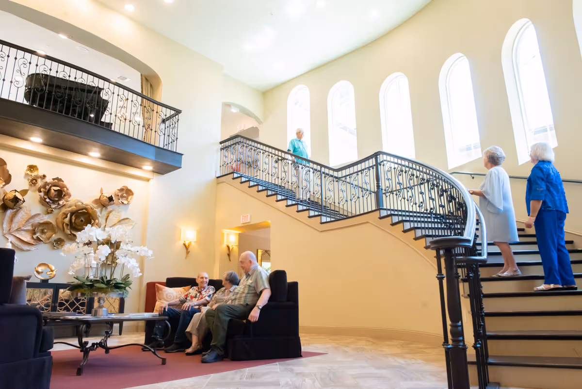 A bright and spacious common area in a senior living facility with a curved staircase and large arched windows letting in natural light. Several elderly people are sitting on sofas and chairs, while others are walking up the stairs. The room is decorated with large floral wall art and a table with a flower arrangement.