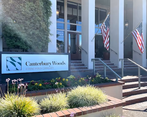 Front entrance of Canterbury Woods with a sign, brick planters, steps up to glass doors, and American flags.