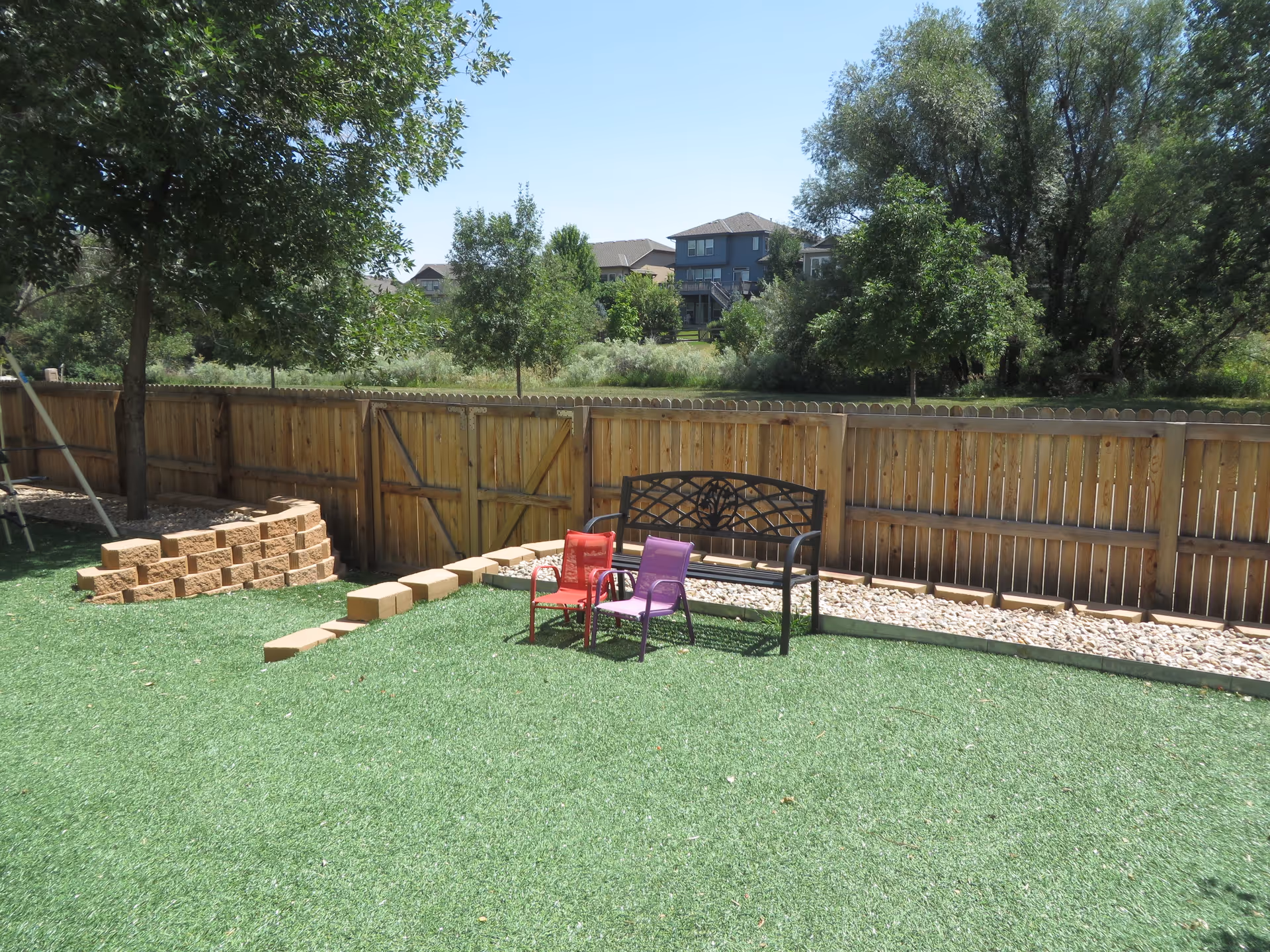 A fenced outdoor area with artificial grass, a wooden fence, a black metal bench, and two small colorful chairs (one red and one purple). There are trees and houses visible in the background under a clear blue sky.