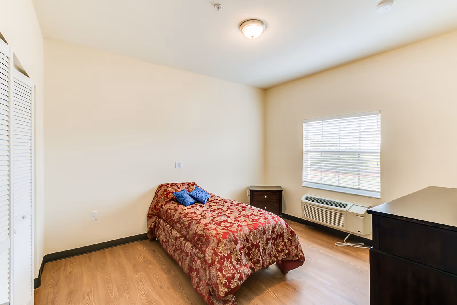 A simple bedroom in Cedar Creek Assisted Living featuring a single bed with a red and gold floral bedspread and two blue pillows. The room has light beige walls, a wooden floor, a window with white blinds, a dark wooden nightstand, a matching dresser, and a white air conditioning unit below the window.