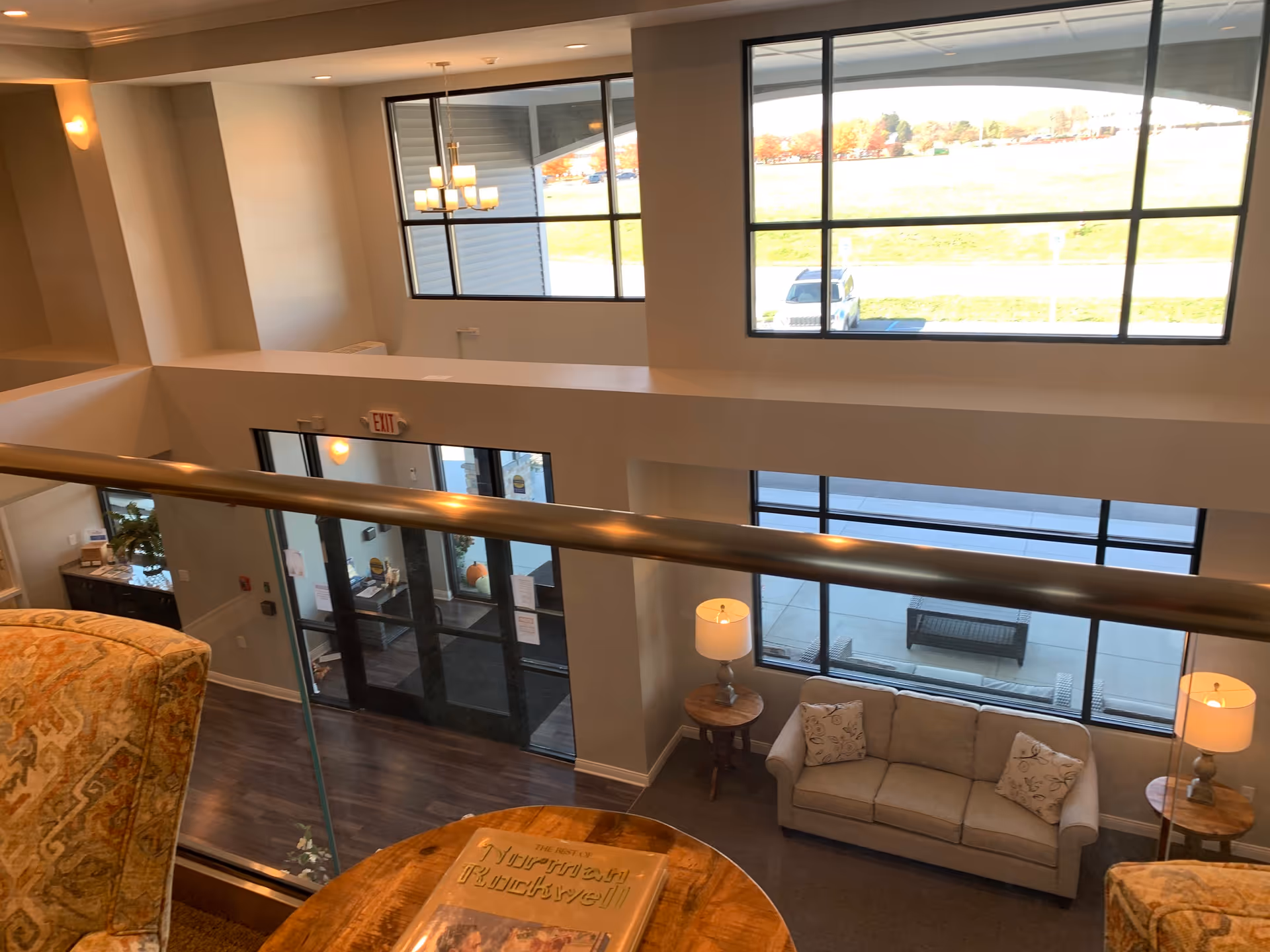 Interior view of a senior living community lobby area taken from an upper level. The space features large windows letting in natural light, a beige couch with two patterned pillows, two side tables with lamps, a wooden round table with a book on it, and a patterned armchair. Outside the windows, a parking lot and grassy area are visible.