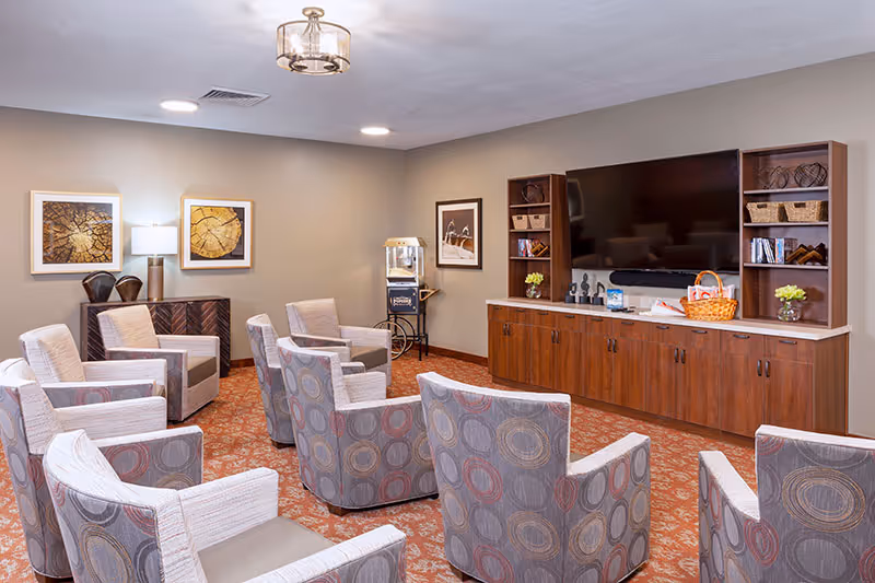 A cozy senior living lounge with upholstered chairs arranged facing a large wall-mounted TV and wooden cabinets.