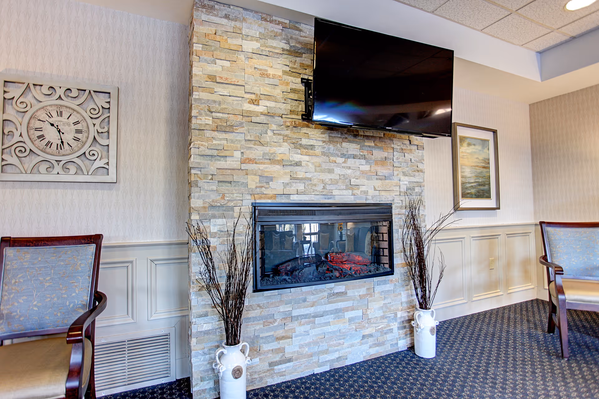 A cozy interior space featuring a stone fireplace with an electric fire, a flat-screen TV mounted above it, two decorative vases with tall dried branches on either side of the fireplace, two upholstered chairs with wooden arms, a decorative wall clock on the left wall, and a framed painting on the right wall.