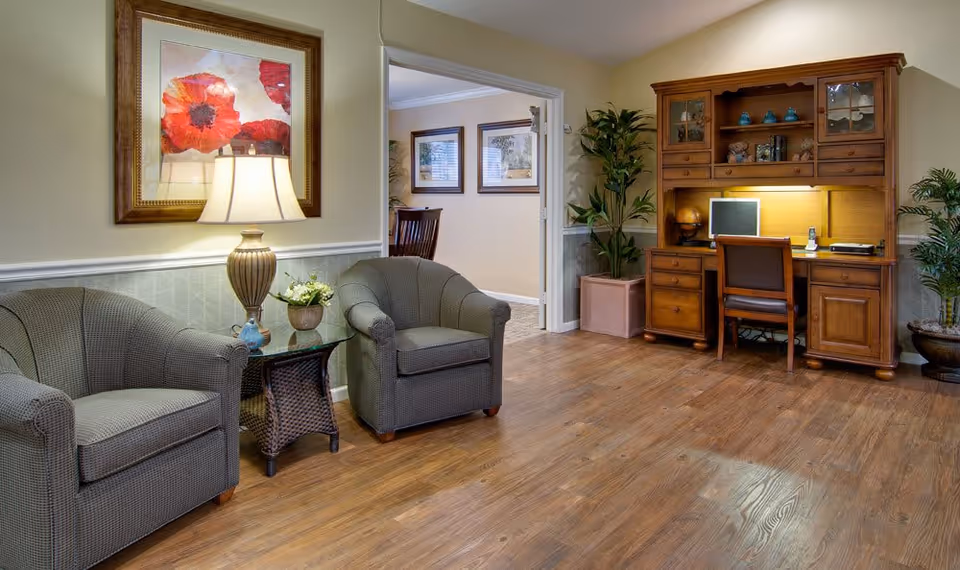 A cozy interior room with two gray upholstered armchairs separated by a glass-top side table holding a lamp and a small plant. On the wall above the chairs is a framed painting of red flowers. To the right, there is a wooden desk with a hutch, a chair, a computer monitor, and some decorative items. The room has wooden flooring and light-colored walls with white trim. A doorway leads to another room with framed pictures on the wall and a wooden chair visible.