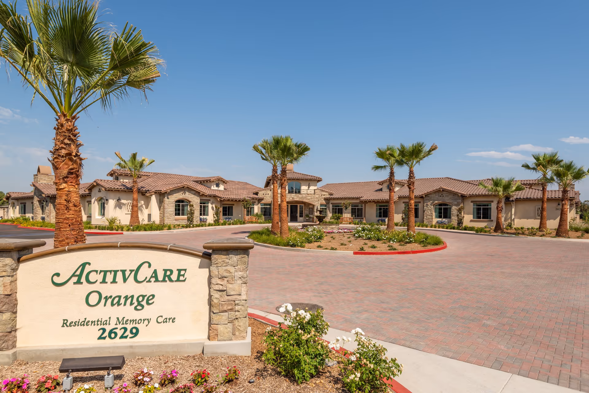 Entrance and front facade of the ActivCare Orange residential memory care facility with a sign, palm trees, and a circular driveway.