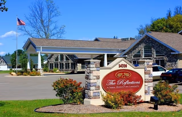 Exterior view of Reflections Memory Care facility showing a stone building with a covered entrance, an American flag on a flagpole, and a landscaped area with bushes and a sign that reads 'The Reflections A Senior Memory Care Community' with the address number 14316.