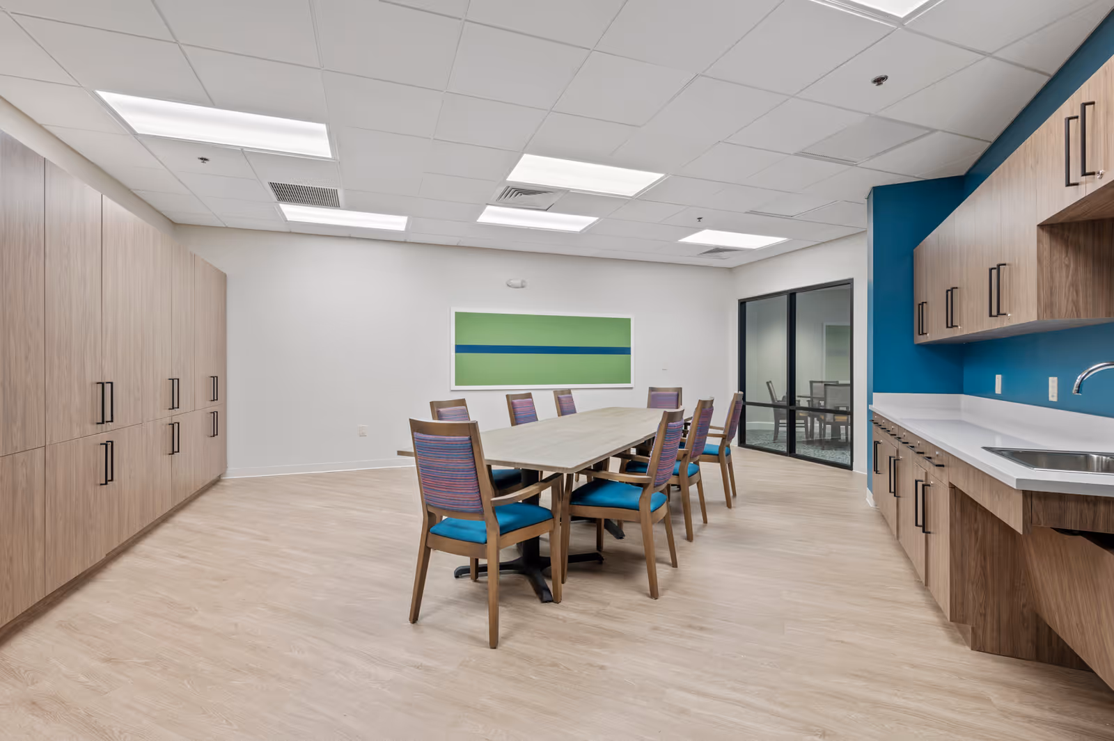 A modern meeting or dining room with a long rectangular table surrounded by eight wooden chairs with blue cushions. The room features light wood flooring, a wall of wooden cabinets on the left, and a kitchenette area with a sink and upper and lower cabinets on the right. The walls are white with one accent wall painted blue, and there is a large green and blue abstract painting on the far wall. The ceiling has recessed lighting panels, and there is a glass door leading to another room.