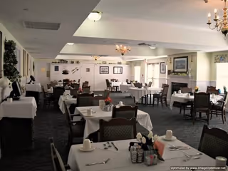 A spacious dining room with multiple tables covered in white tablecloths, each set with cups, saucers, and silverware. The room features chandeliers, framed artwork on the walls, and large windows allowing natural light to enter. Chairs are neatly arranged around the tables, and there is a fireplace visible on the right side of the room.