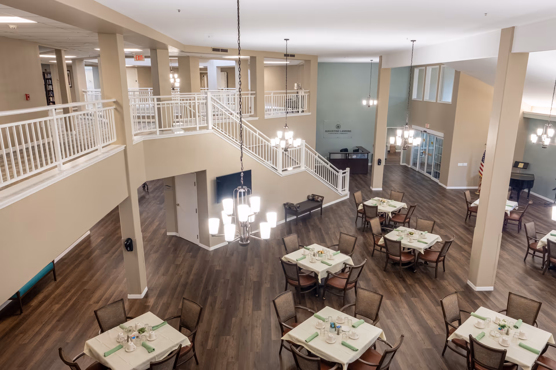 Spacious dining area in Augustine Landing facility with multiple tables set for meals, wooden flooring, beige walls, and modern hanging light fixtures. There is a staircase leading to an upper level with white railings and a reception desk visible in the background.