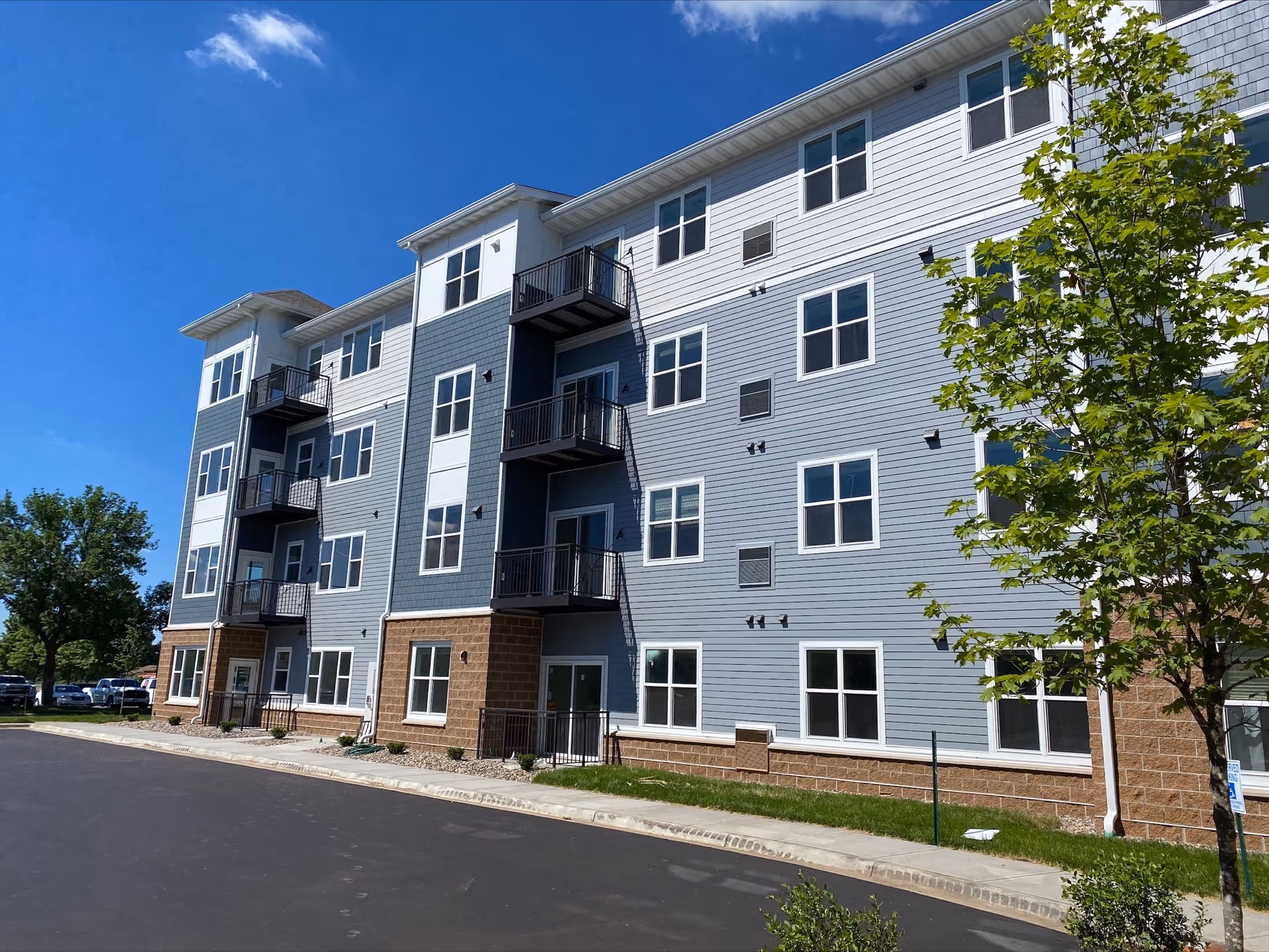 Exterior view of a modern four-story senior living facility building with multiple windows and small balconies, surrounded by a paved driveway, green grass, and trees under a clear blue sky.