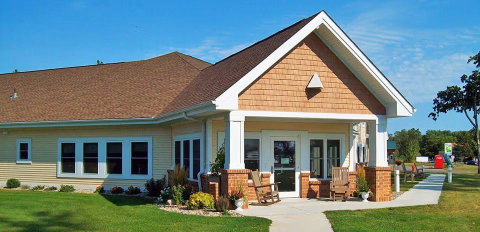 Exterior view of a single-story building with beige siding and a brown shingled roof. The entrance features a covered porch supported by white columns and brick bases, with two wooden rocking chairs on the porch. The building is surrounded by a well-maintained lawn and landscaping, with a clear blue sky overhead.