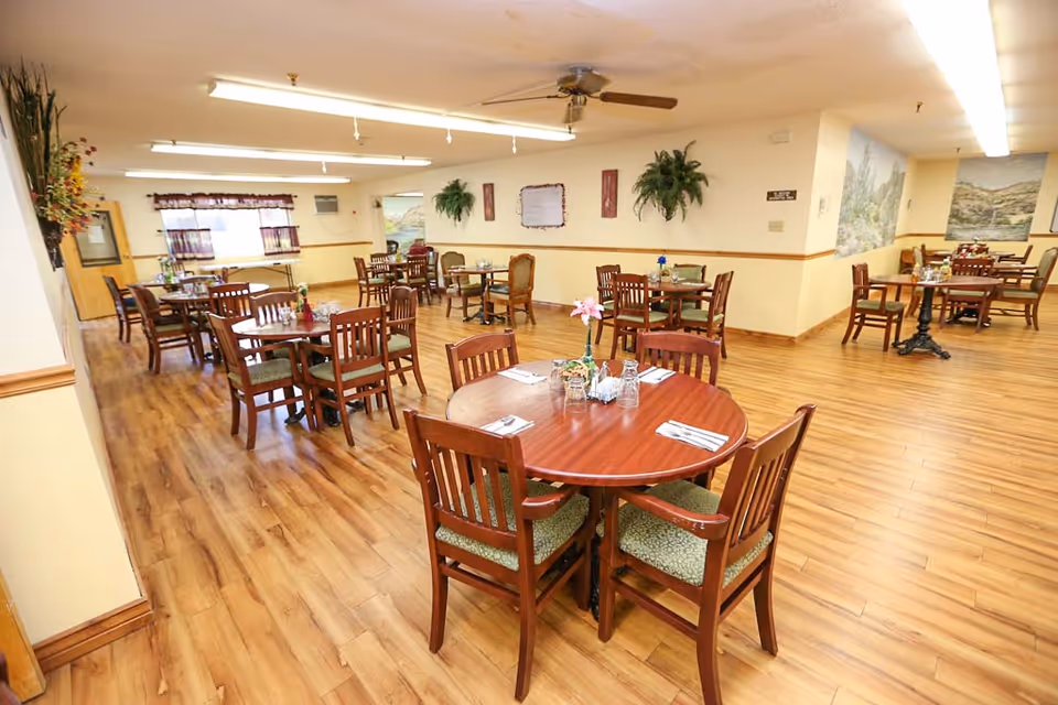 A spacious dining room with multiple wooden tables and chairs arranged neatly on a polished wooden floor. The room is well-lit with fluorescent ceiling lights and has ceiling fans. There are decorative plants on the walls and framed artwork, with windows covered by curtains in the background.