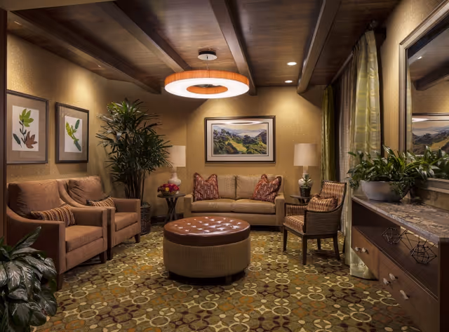 A cozy living room with warm lighting featuring a beige sofa with patterned cushions, two matching armchairs, a round ottoman in the center, a wooden side table with a lamp and flowers, framed artwork on the walls, a large plant in the corner, and a wooden console with decorative items and a large mirror above it.