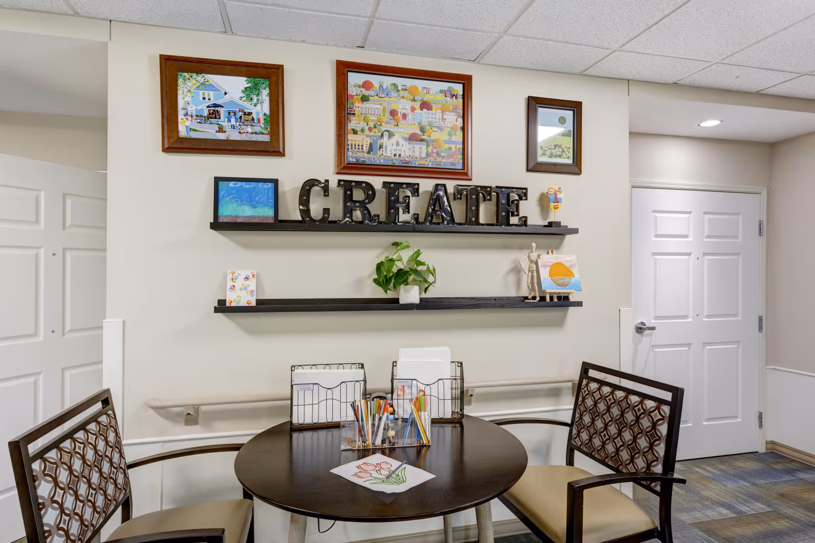 Small craft/activity area with a round table and chairs beneath wall shelves holding framed art and decorative letters spelling "CREATE".
