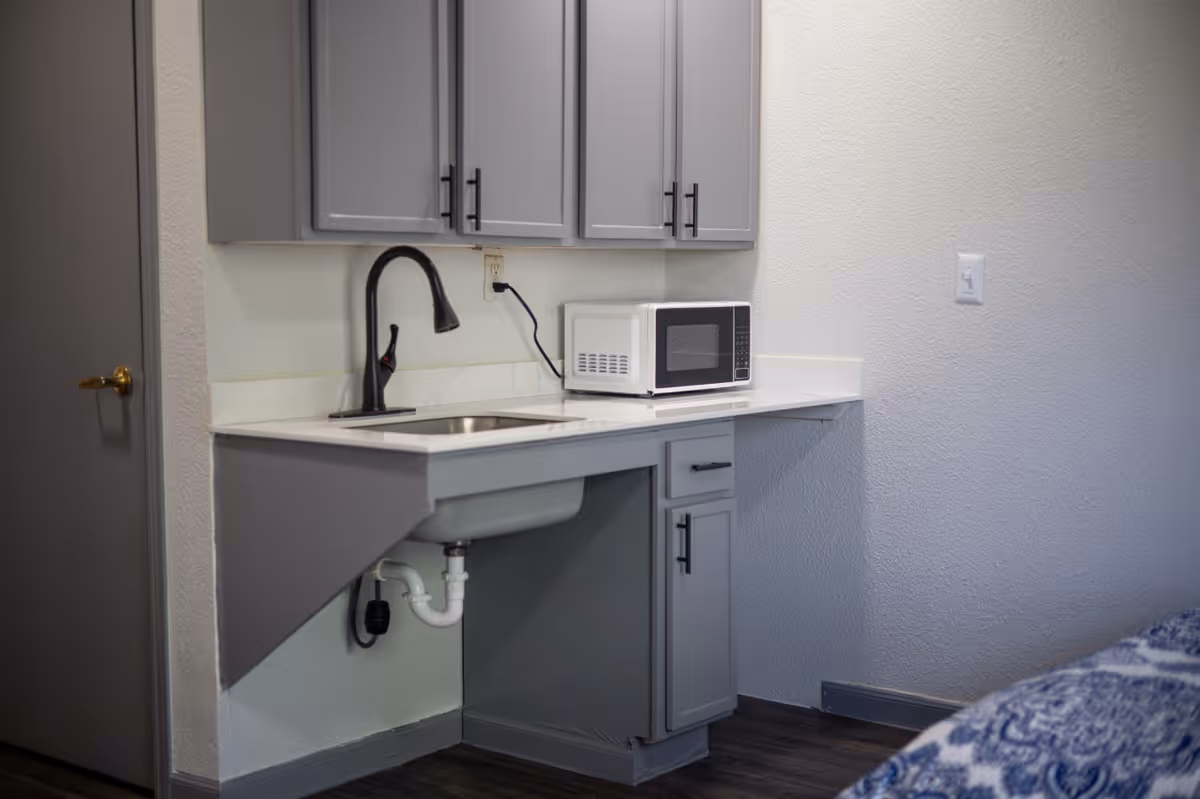Compact kitchenette with gray cabinets, a countertop sink, and a microwave in a senior living unit.